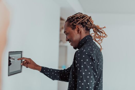 Smiling African American man using modern smart home system, controller on wall, positive young man switching temperature on thermostat or activating security alarm in apartment. High quality photo