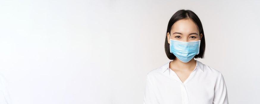 Covid and healthcare concept. Close up portrait of asian woman, office lady in face mask, smiling, using protection from coronavirus during pandemic, white background.