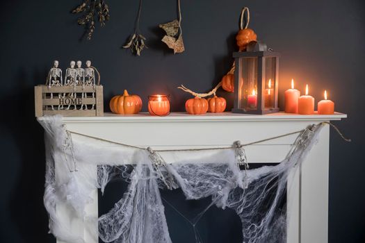 Halloween home decoration. Plastic toy skeletons in a wooden box on a fireplace against a dark blue wall. A garland of skeletons. Cobweb on the dresser. Orange candles and lantern.
