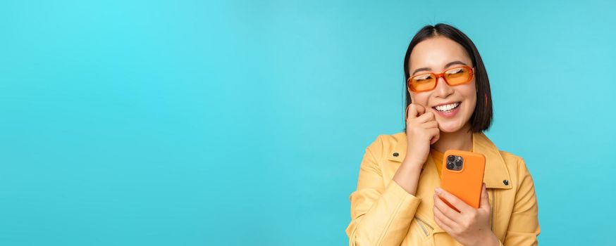 Happy stylish asian girl using smartphone and laughing, smiling at camera, standing over blue background. Copy space