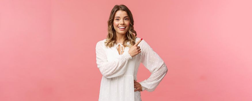 Upbeat good-looking caucasian blond girl in white dress, pointing upper right corner and smiling at camera with interested, curious face discuss special promo spring discounts, pink background.