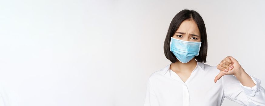 Portrait of asian woman in medical face mask showing thumbs down with disappointed, tired face expression, standing over white background.