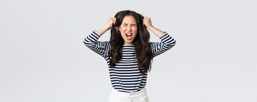 Lifestyle, people emotions and casual concept. Pissed-off mad and angry asian young woman tossing hair, pulling it from head with screams and closed eyes, standing bothered white background.