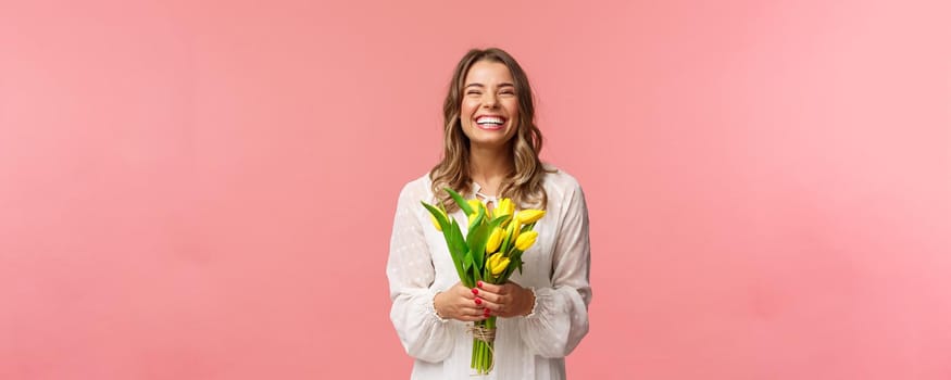 Holidays, beauty and spring concept. Portrait of happy excited charming blond girl receive flowers, buying yellow tulips herself, smiling and laughing joyfully, stand pink background.