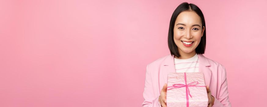 Smiling asian businesswoman in suit, giving you gift in wrapped box, standing over pink background. Copy space