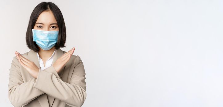 Coronavirus and workplace concept. Image of asian businesswoman, female in medical face mask shows stop, cross prohibit gesture, stands over white background.