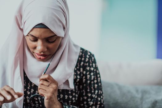 African woman using smartphone while sitting on the sofa at home wearing hijab clothes. High-quality photo