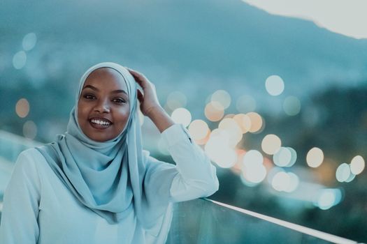 African Muslim woman in the night on a balcony smiling at the camera with city bokeh lights in the background. High-quality photo