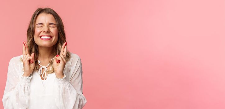 Portrait of excited hopeful blond girl making wish crossed fingers for good luck, close eyes and smiling putting all effort into pray, pleading for dream come true, anticipating over pink background.