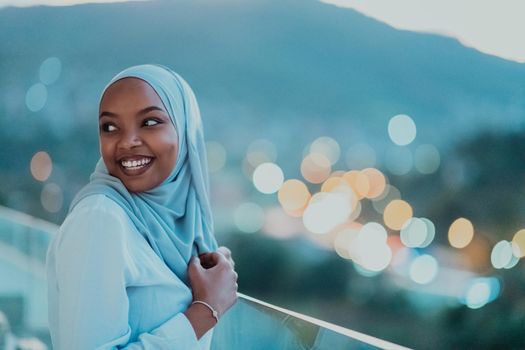 African Muslim woman in the night on a balcony smiling at the camera with city bokeh lights in the background. High-quality photo