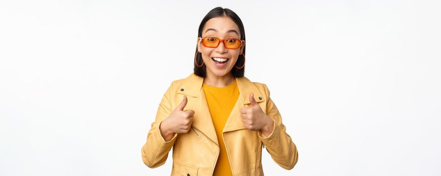 Enthusiastic asian girl smiles, shows thumbs up and looks happy, saying yes, approve and recommend smth, standing over white background.
