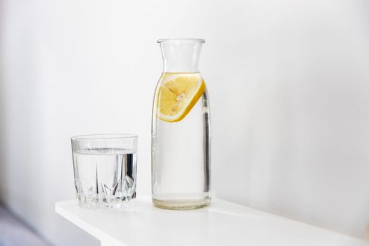 A refreshing glass of water, a bottle with a slice of lemon inside is on the dresser in the summer heat