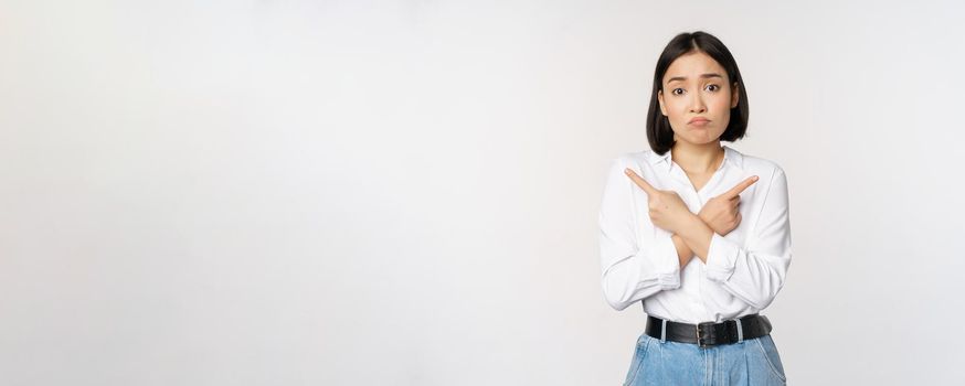 Confused sad asian girl cant decide, pointing fingers left and right, sulking and shrugging indecisive, cannot choose, standing over white background.