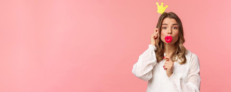 Spring, happiness and celebration concept. Close-up portrait of funny and cute, silly blond girl in white dress, holding big lips mask and crown, partying and having fun, pink background.