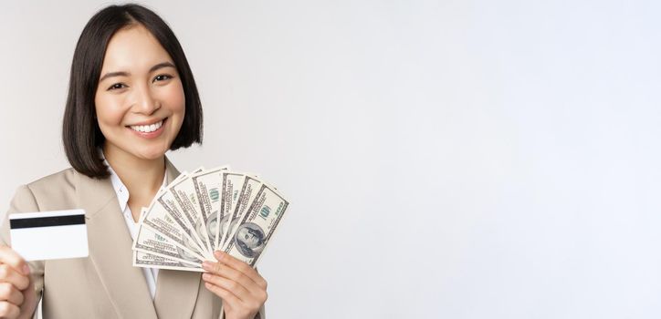 Close up of asian businesswoman, office lady showing credit card and money dollars, standing in suit over white background.