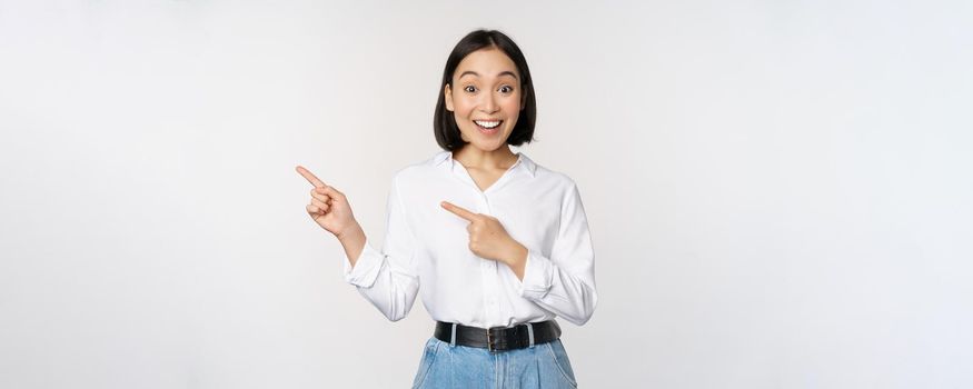 Portrait of excited young asian woman, office lady pointing fingers left at discount, showing sale banner, standing over white background.