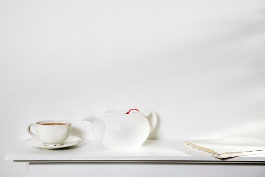White on white. A cup of tea, a kettle, and a notebook stand on a white panel of an artificial fireplace. Empty space.