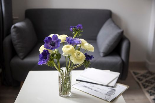 Bouquet of white ranunculus and blue anemone in the vase on a white coffee table near sofa with plaid. The books and journal. Shadow