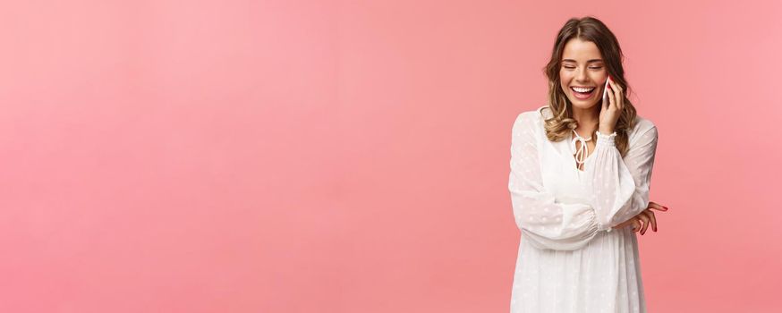 Communication and technology concept. Attractive caucasian female in white dress, blond short haircut, hold smartphone, talking on mobile phone and laughing, smiling carefree, pink background.