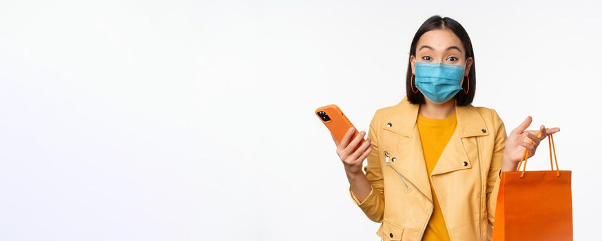 Image of stylish asian girl shopper, holding smartphone and shopping bag without store logo, wearing medical face mask from covid-19, white background.