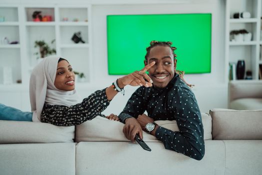 African Couple Sitting On Sofa Watching TV Together Chroma Green Screen Woman Wearing Islamic Hijab Clothes. High quality photo