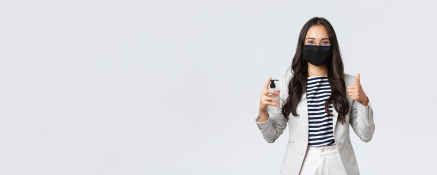 Business, finance and employment, covid-19 preventing virus and social distancing concept. Smiling businesswoman in face mask showing hand sanitizer and thumbs-up, ask to use it.