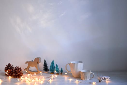 Christmas composition on the table. Wooden gray rocking horse, four small artificial snow-covered trees, angel, garland and mugs