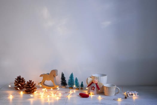 Christmas composition on the table. Wooden gray rocking horse, four small artificial snow-covered trees, angel, garland and mugs