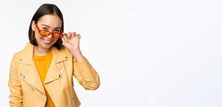 Beautiful asian girl in sunglasses, smiling and looking happy, posing in yellow jacket against white background. Copy space
