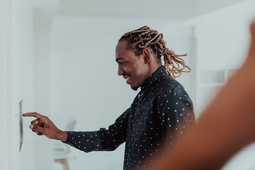 Smiling African American man using modern smart home system, controller on wall, positive young man switching temperature on thermostat or activating security alarm in apartment. High quality photo