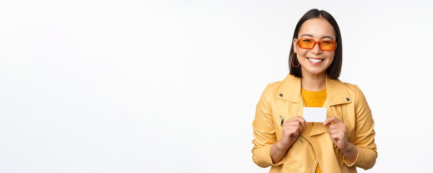 Portrait of beautiful modern asian girl in sunglasses, smiling happy, showing credit card, standing over white background. copy space