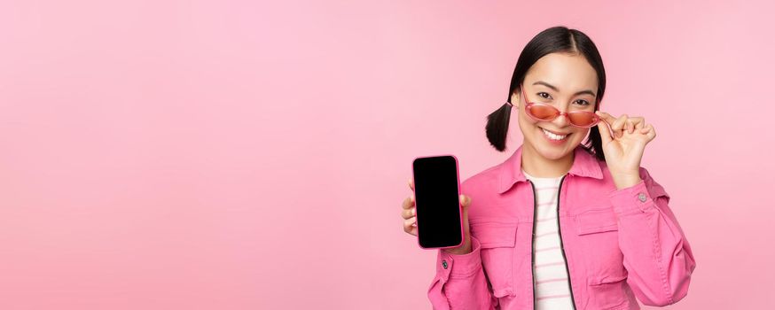 Portrait of stylish modern asian girl shows mobile phone screen, smartphone app interface, standing in sunglasses against pink background.