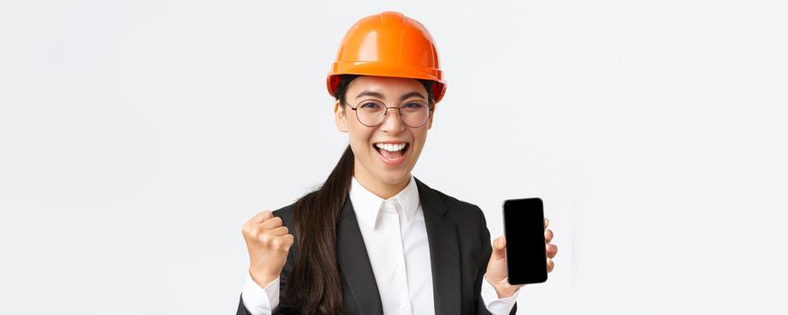 Successful winning asian female engineer, architect in safety helmet and suit, showing smartphone screen, fist pump and shouting yes from rejoice, triumphing over achievement, white background.