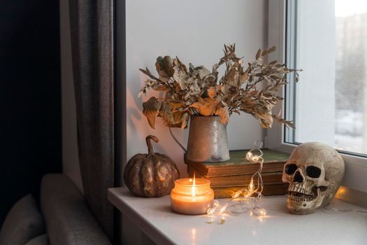 Bouquet of dried autumn leaves in a zinc jug, plastic skull, artificial gilded pumpkin, vintage books decorate the window for Halloween