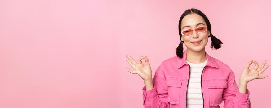 Mindfulness and wellness concept. Smiling korean girl in stylish outfit meditating, listening mantra, holds hands in zen, peace pose, practice yoga, standing over pink background.