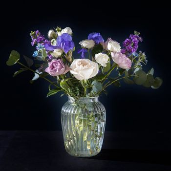 Bouquet of hackelia velutina, purple and white roses, small tea roses, matthiola incana and blue iris in glass vase . Dark blue wall