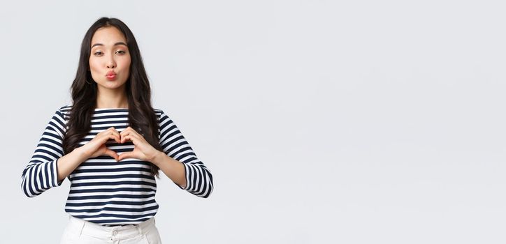 Lifestyle, people emotions and casual concept. Lovely smiling adorable asian woman showing heart sign and smiling, express sympathy or care, sending air kiss at camera, standing white background.