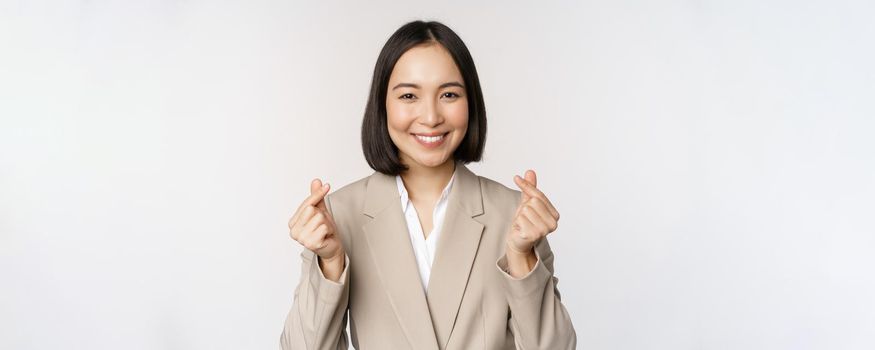 Cheerful asian saleswoman, smiling and showing finger hearts sign, standing in suit over white background.