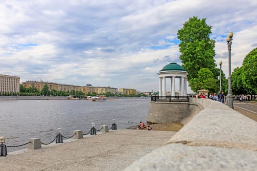 Moscow, Russia - May 15, 2021: View of Pushkin embankment. People sits on log by the river. ship sails under the bridge. Mother and child stand by the steps. Gorky Central Park of Culture and Leisure