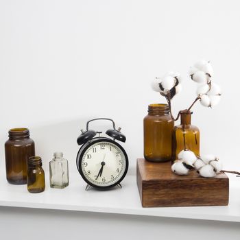 Cotton flowers in glass brown small pharmaceutical bottles on white table surface as room decoration. The clock shows six.
