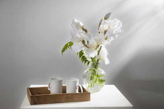 A bouquet of three white irises and a fern in a transparent vase on the table. Two ceramic tea in the tray. Breakfast