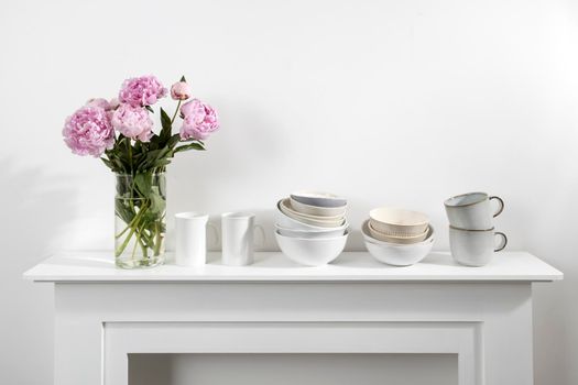 Bouquet of pink peonies, a stack of cups, plates, bowls, white chest of drawers in the interior of the kitchen