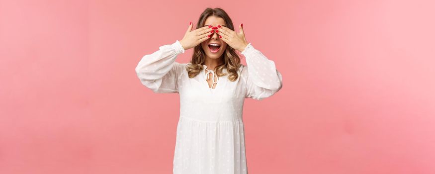 Excited cute blond girl in white spring dress waiting for gifts at surprise party, cover eyes blindfolded standing pink background, open mouth in amazement and anticipation.