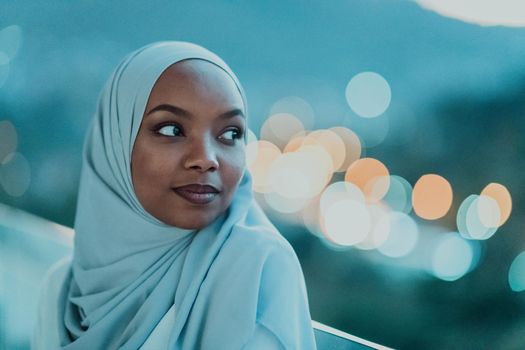 African Muslim woman in the night on a balcony smiling at the camera with city bokeh lights in the background. High-quality photo