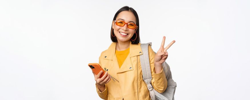 Young asian woman traveller, girl tourist in sunglasses, holding backpack and mobile phone, using application on smartphone, standing over white background.