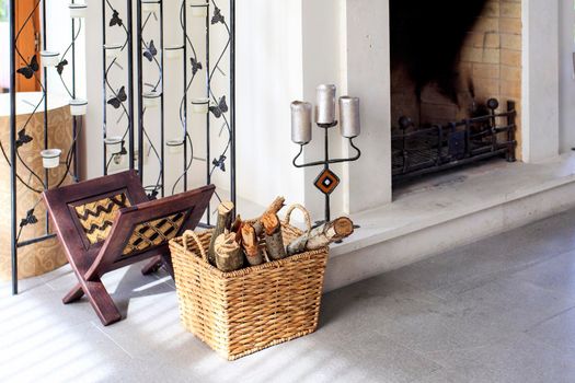 Fragment of a fireplace with a wicker basket with firewood, a stand for magazines and candles in a candlestick. cosiness