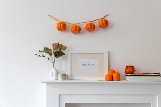 Preparing your home for Halloween. A garland of pumpkins on the wall above fake dresser panel. Frame with the inscription, orange candles and lantern. Corrugated vase with dried eucalyptus branch.