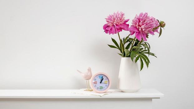 Red, pink peonies in a white vase on a table, clock, and against a white wall background. Wide frame. Copy space.