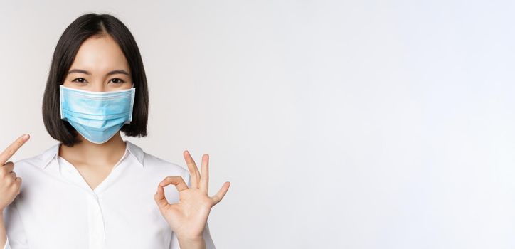 Portrait of asian girl in medical mask showing okay sign and pointing at her covid protection, standing over white background. Copy space