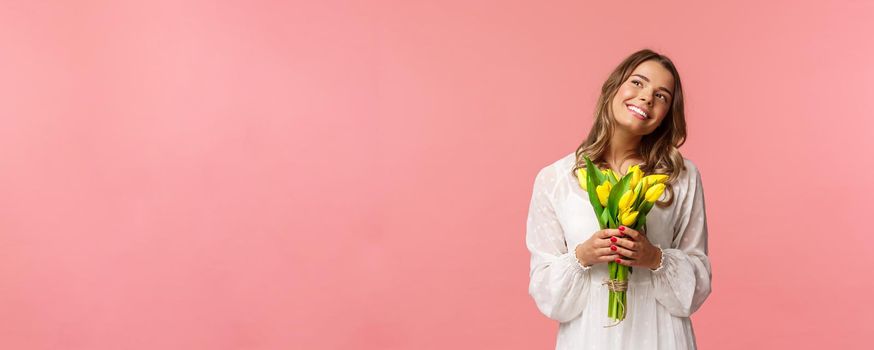 Holidays, beauty and spring concept. Portrait of dreamy, happy blond girl feeling romantic daydreaming about her girlfriend, holding yellow tulips, wear white dress, standing pink background.